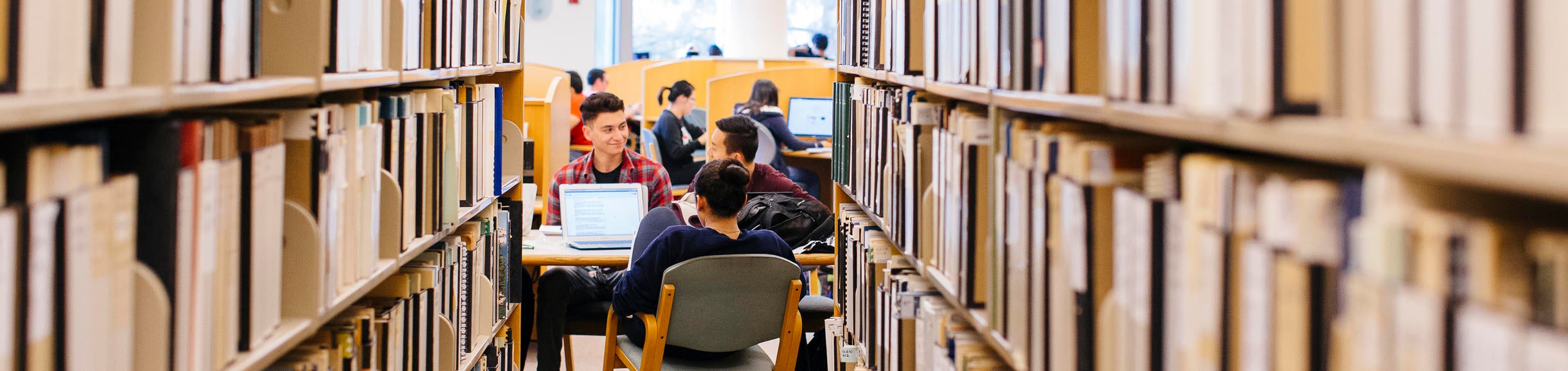 A group of students studies together at a table in the library as seen from between two shelves of books.